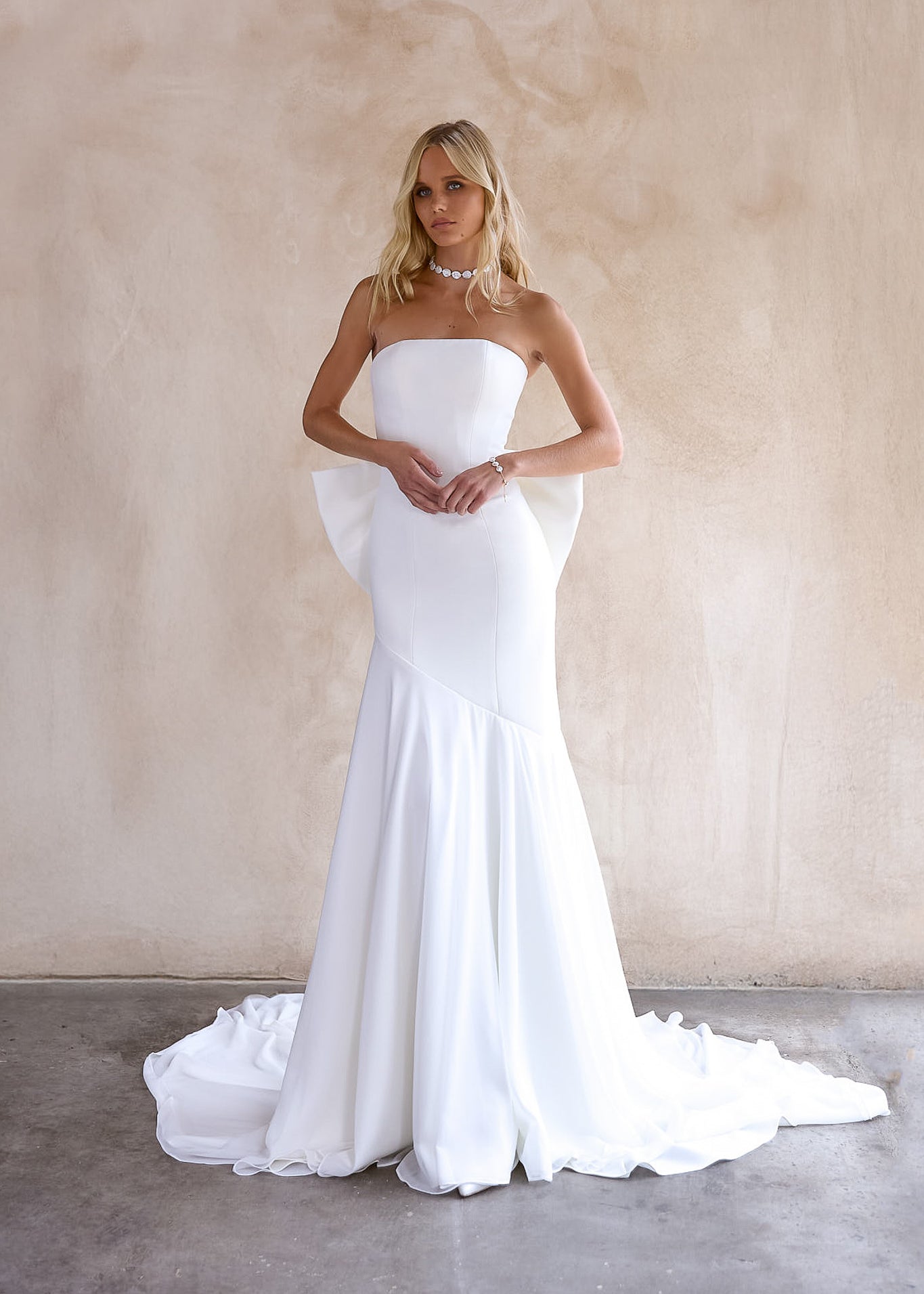 Front Image of the June Wedding dress by The Bryde, featuring a detachable bow and pearl jewellery. Made from crepe main fabric and a soft chiffon angled skirt