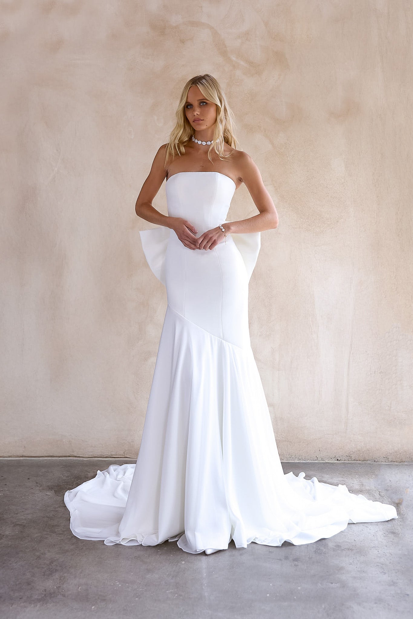 Front Image of the June Wedding dress by The Bryde, featuring a detachable bow and pearl jewellery. Made from crepe main fabric and a soft chiffon angled skirt