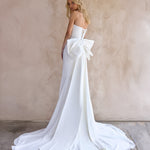 Back Image of the strapless June Wedding dress by The Bryde, featuring a detachable bow and pearl jewellery. Made from crepe main fabric and a soft chiffon angled skirt